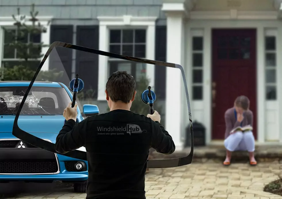 WindshieldHUB technician installing a new windshield on a blue car with a shattered front window, while a woman reads a book on the steps of a house in the background